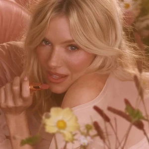 Close-up portrait of Sienna Miller holding a pink lipstick amongst soft floral blooms for the Charlotte Tilbury Pillow Talk in Bloom campaign.