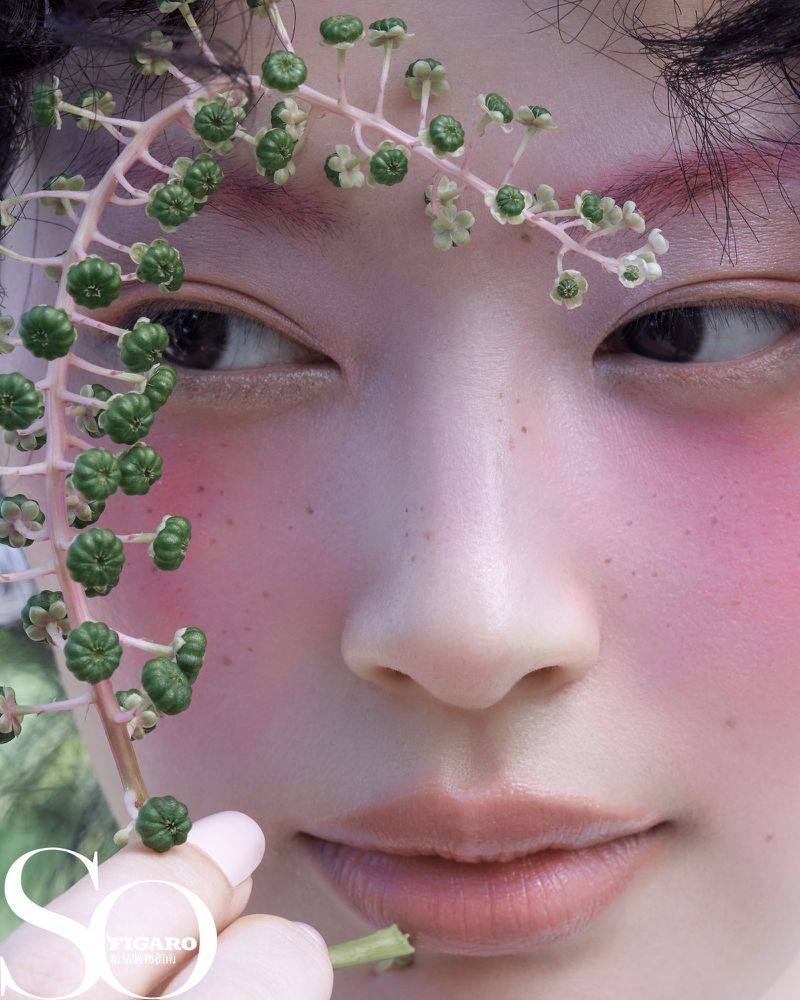 Close-up portrait of a person with fair skin and pink blush, adorned with a flowering vine against an out-of-focus green background.