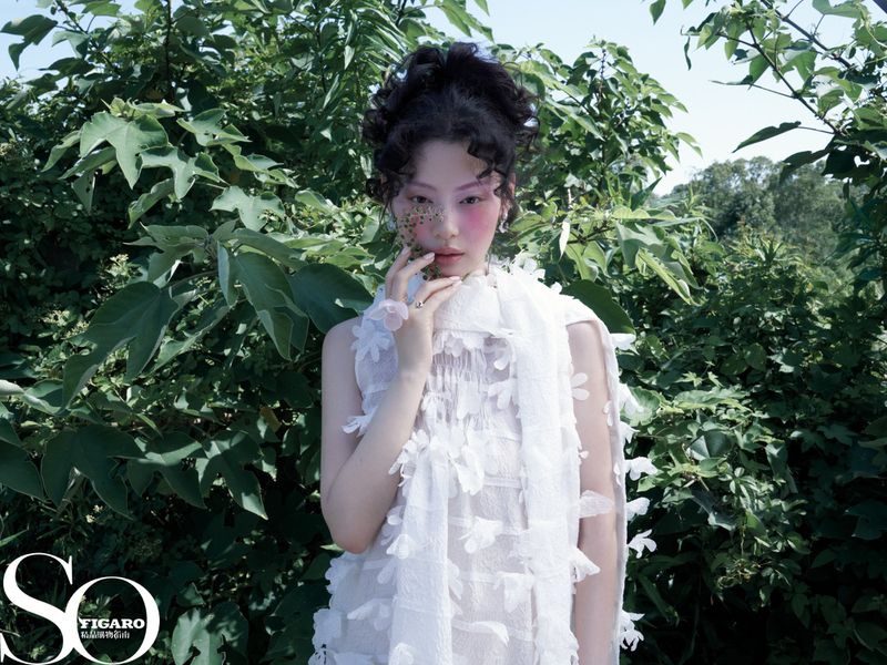 East Asian woman with curly updo and pink blush posing in a textured white appliqué dress against green bushes.