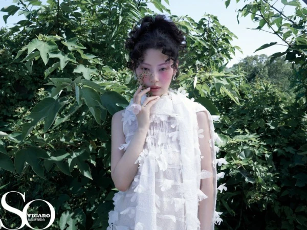 East Asian woman with curly updo and pink blush posing in a textured white appliqué dress against green bushes.
