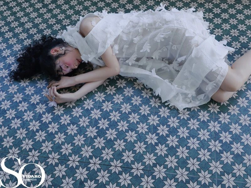 Young woman in a white dress lies on a patterned tile floor, a high-angle view capturing her relaxed pose and the intricate dress details.