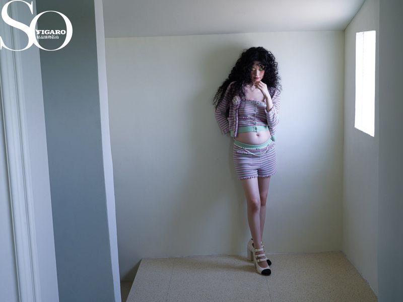 Young woman in a striped, two-piece outfit poses in a corner. The background is a minimalist photo studio space. She wears heels.
