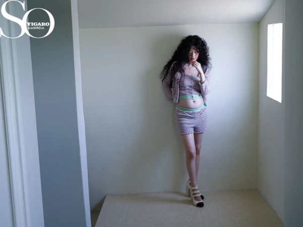 Young woman in a striped, two-piece outfit poses in a corner. The background is a minimalist photo studio space. She wears heels.