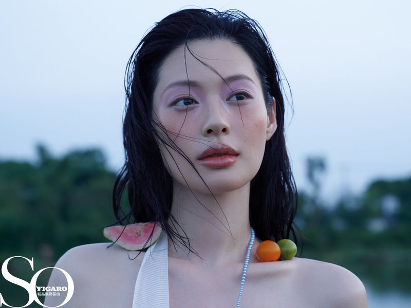 Close-up portrait of a woman with fruit accessories and pink eye shadow, outdoors, for a fashion magazine cover.