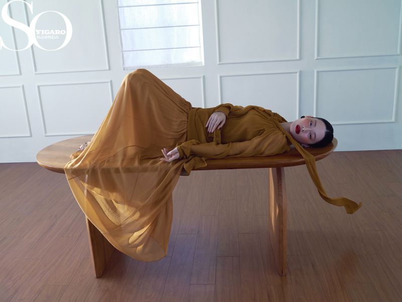 Elegant woman in flowing amber gown poses gracefully on a wooden table in a minimalist indoor setting with soft, diffused natural light.