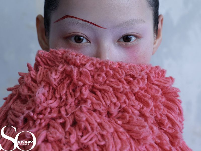 Close-up shot of a woman with creative makeup, including a drawn-on eyebrow, covering her face with a fluffy, textured garment.
