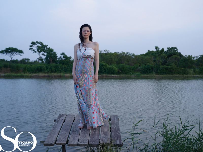 A woman in a dress stands on a wooden dock by a lake surrounded by trees, in an outdoor setting, captured in a medium shot.