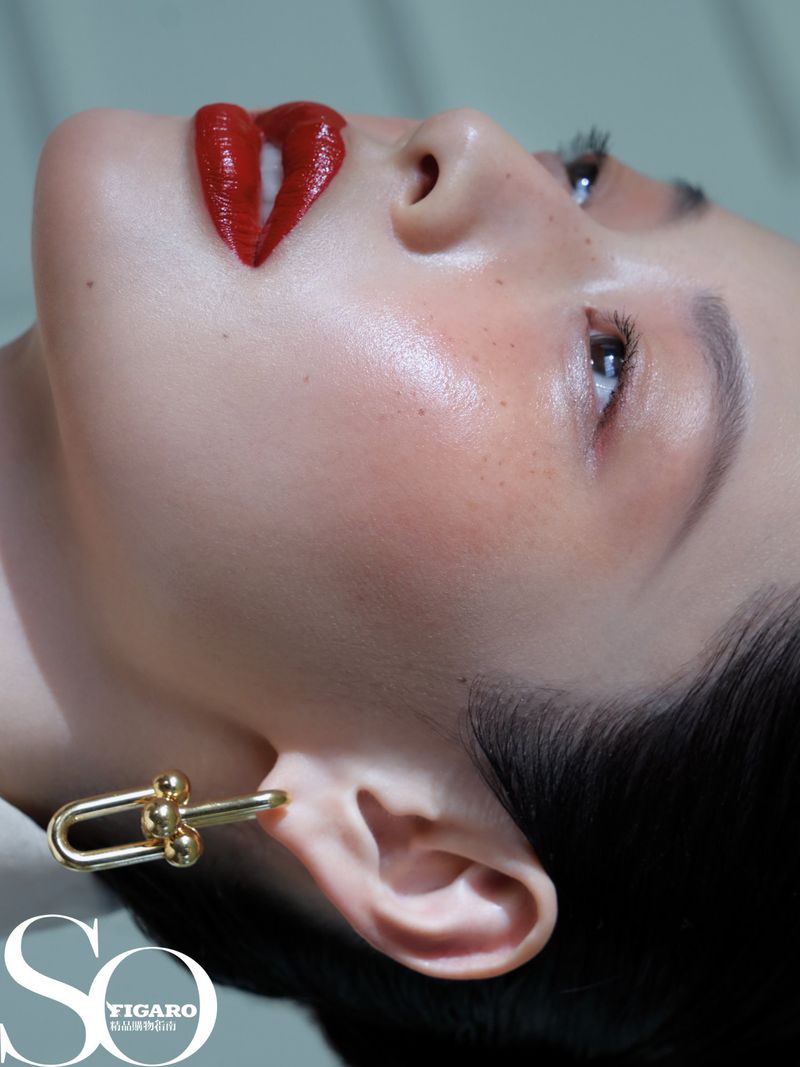 Close-up studio shot of a woman with red lipstick shaped like a heart, a gold earring, light skin, and freckles, shot at eye level.
