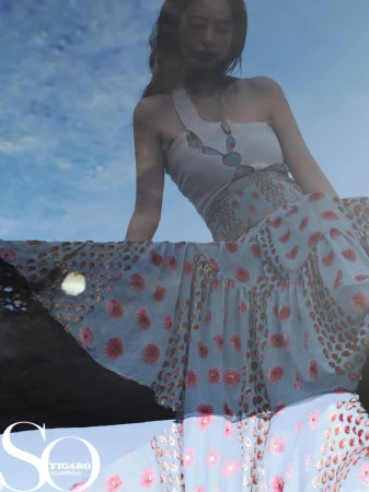 A low-angle shot of a young woman in a floral dress, sitting on a ledge with a blue sky and clouds in the background.
