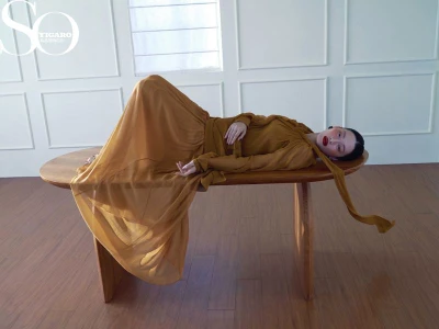 Model in a flowing mustard yellow gown reclining on a wooden bench against a white paneled wall with soft light.