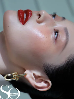 Close-up beauty profile of an East Asian woman with glossy red lipstick, light freckles, and a sculptural gold earring.