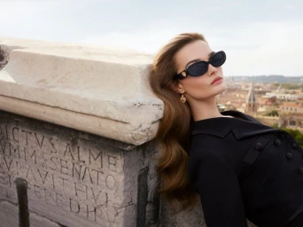 Model Lulu Tenney wearing black sunglasses and black attire leaning against an inscribed stone wall for Dolce & Gabbana Devotion.
