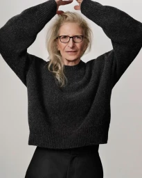 Annie Leibovitz wearing a dark grey knit sweater and black trousers with glasses against a white background