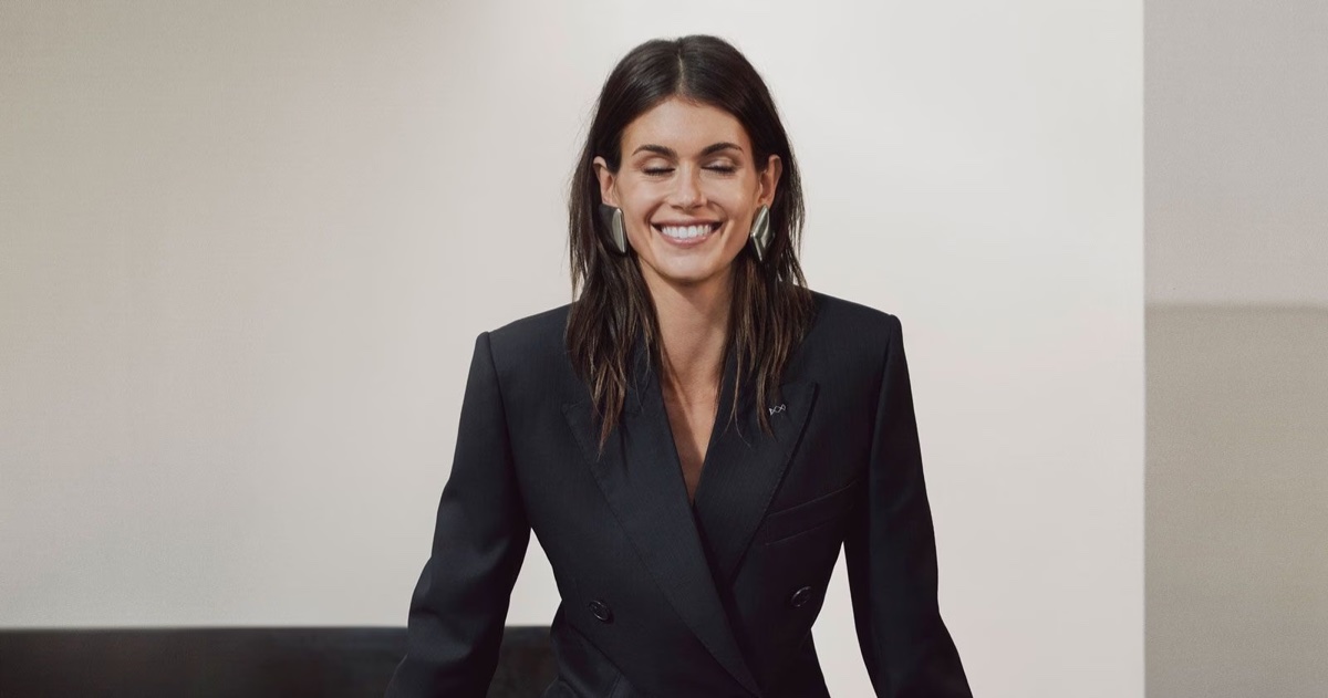 Model Kaia Gerber smiling in a black blazer with large silver earrings against a neutral background for Givenchy