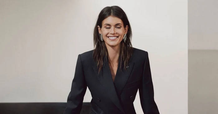 Model Kaia Gerber smiling in a black blazer with large silver earrings against a neutral background for Givenchy