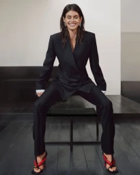 Woman in a black blazer and matching trousers seated on a black chair against a minimalist neutral studio background.