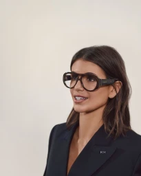 Portrait of a woman wearing oversized tortoiseshell glasses and a dark blazer against a neutral background