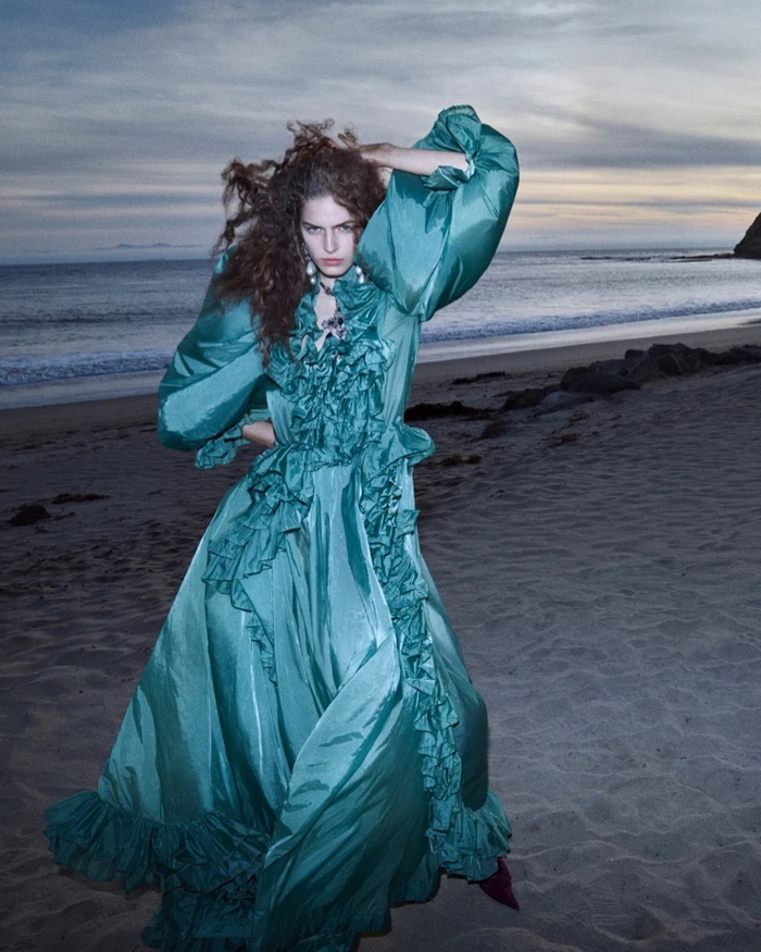 Model in teal ruffled silk dress posing on a sandy beach at dusk with ocean waves and a cloudy horizon.