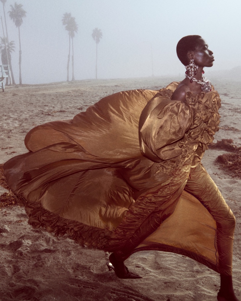 Model in a voluminous bronze gown walking on a hazy sandy beach with palm trees in the background