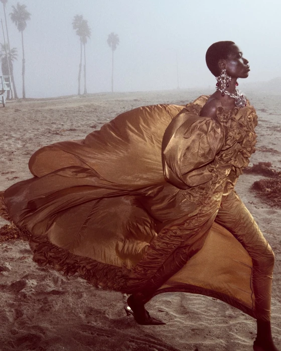 Model in a voluminous bronze gown walking on a hazy sandy beach with palm trees in the background