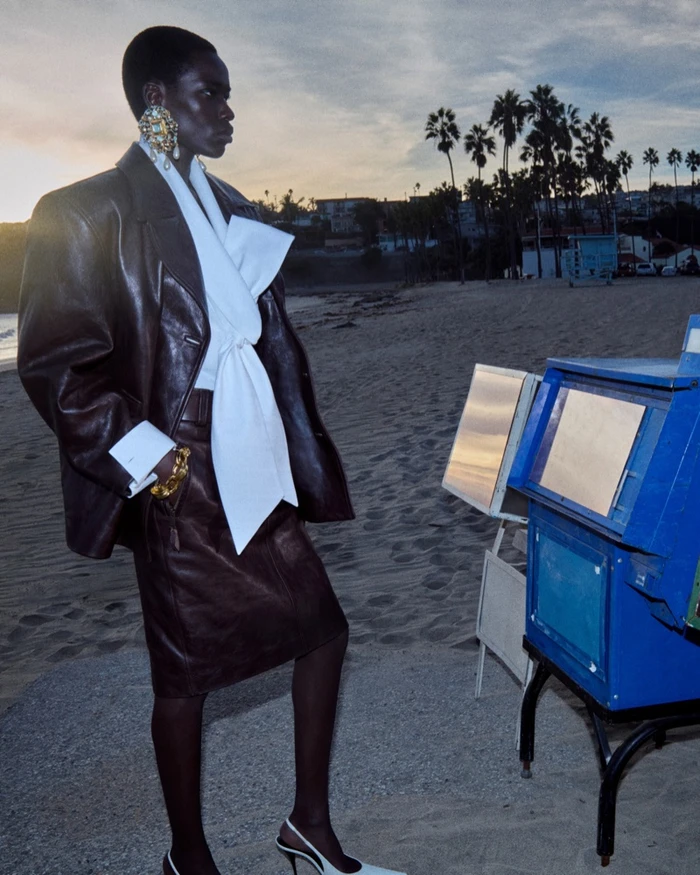 Model standing on a sandy beach wearing a dark brown leather blazer and white draped blouse against a sunset sky.