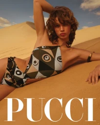 Model lying on sand dunes wearing patterned one-piece swimsuit and gold jewelry under a clear sky for Emilio Pucci.
