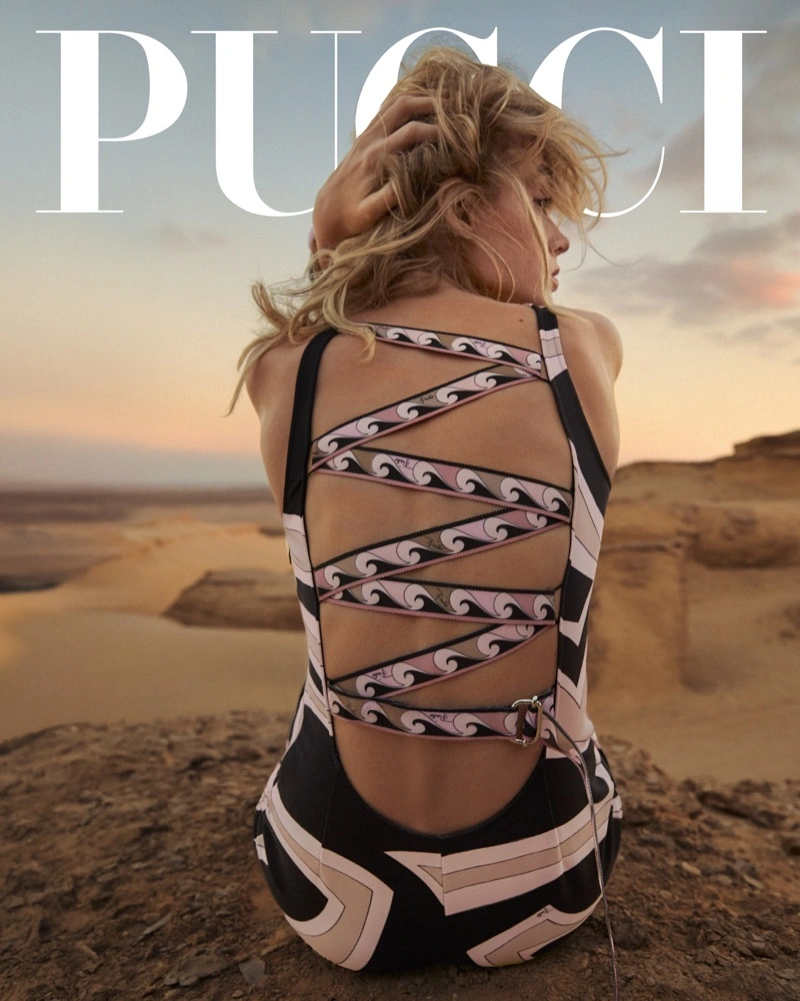 Model seated on rocky terrain wearing a patterned swimsuit with crisscross straps, seen from the back against a desert sunset background.