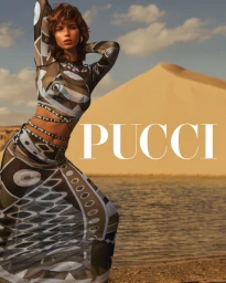 Model wearing patterned sheer top and skirt posing by a sandy dune and water under a cloudy sky.