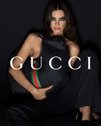 Emily Ratajkowski holding a black Gucci handbag wearing dark trousers against a dark patterned background