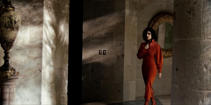 A woman in an orange dress walking through a stone hallway with a decorative vase and a round mirror.