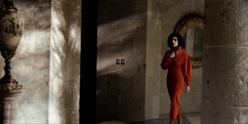 A woman in an orange dress walking through a stone hallway with a decorative vase and a round mirror.