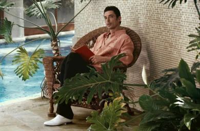 Alberto Guerra seated in a wicker chair by a pool wearing a peach shirt and white shoes