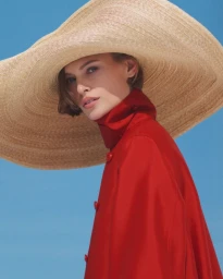 Model wearing a large straw sun hat and a red coat against a clear blue sky