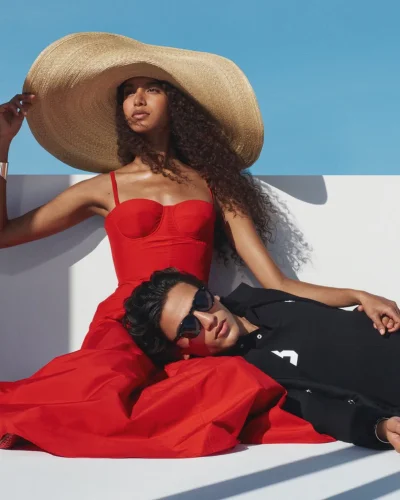 Model in wide-brimmed straw hat and red dress with man resting head on lap under blue sky