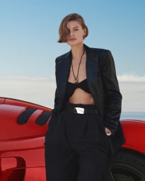 Model leaning against a red car wearing a black blazer and black trousers under a clear sky.