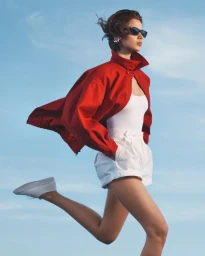 Model wearing a red jacket and white shorts jumping against a bright blue sky background.