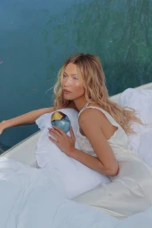 Bella Hadid holding an Orebella perfume bottle while reclining on white pillows beside water for Jasmine Blues