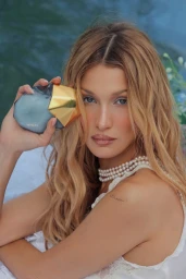 Bella Hadid holding an Orebella perfume bottle while wearing layered pearl necklaces and a white top against a soft blue background