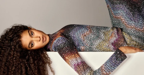 Model Luna Yohannan reclining in a shimmering multicolored patterned knitwear outfit against a grey studio backdrop.