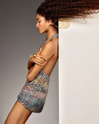 Model wearing a sequined minidress leaning against a white panel with voluminous curly hair blowing horizontally
