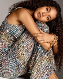 Model with long dark curly hair wearing a sequined outfit and gold chain bracelet against a neutral background