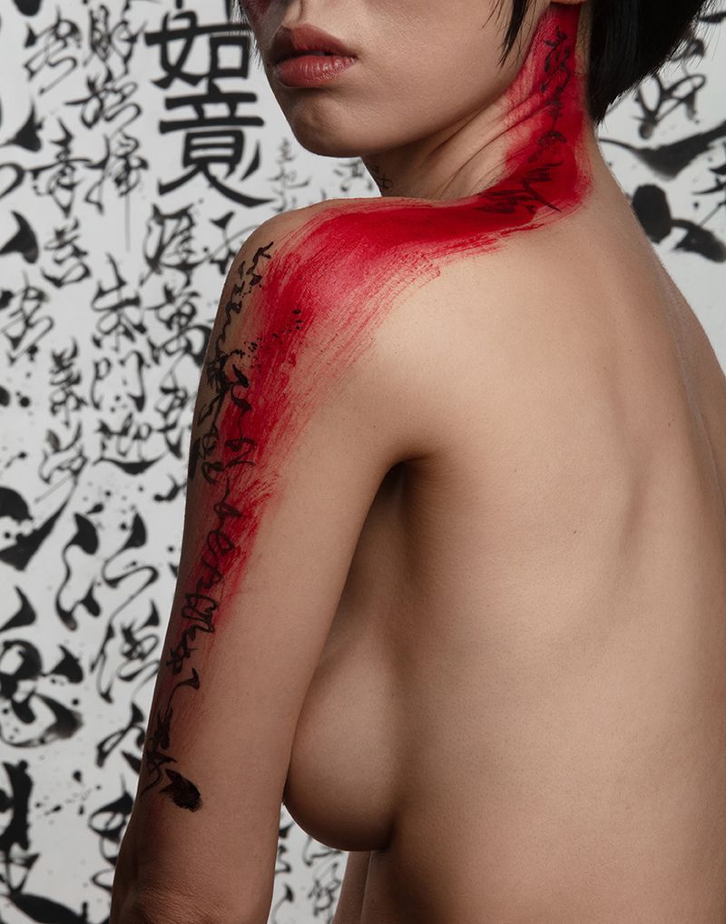 Artistic shot of a young woman with red body paint on her arm and neck, posing against a backdrop with handwritten text.