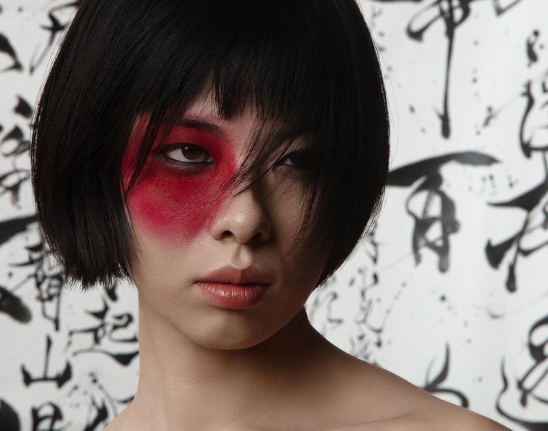 Close-up portrait of an Asian woman with dramatic red makeup on one side of her face and short, straight black hair, set against a background of black calligraphic art.