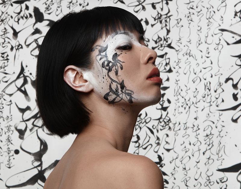 Profile shot of an adult female with black bob haircut and face paint, against a black and white background with calligraphic designs.