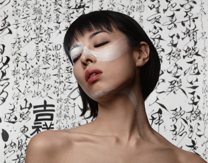 Portrait of an Asian woman with white makeup covering her eyes and black bob haircut posing in front of a calligraphy background.
