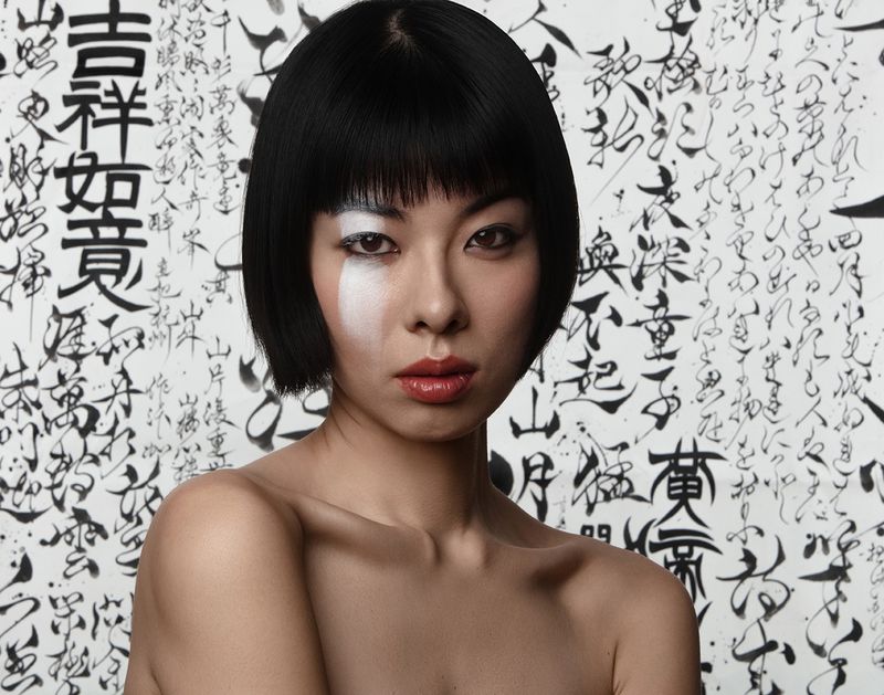 Asian woman with blunt bob hairstyle and artistic white makeup stands against a backdrop of bold black Japanese calligraphy.