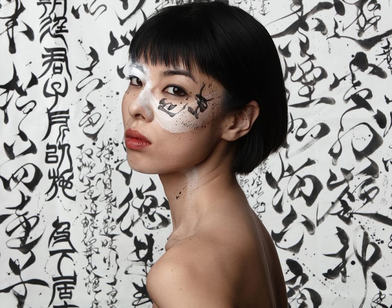 Portrait of an Asian woman with artistic makeup in black and white tones against a backdrop of Asian calligraphy characters.