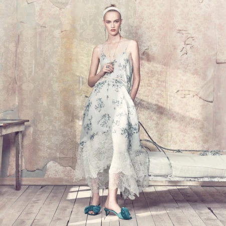 Model wearing a floral lace-trimmed slip dress, white headband, and green bow mules standing in a rustic room.