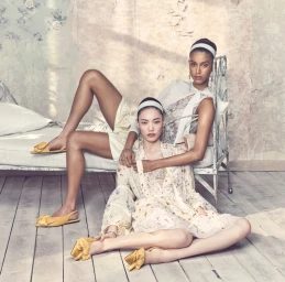 Two female models in pale patterned dresses and yellow shoes posing on a rustic metal bed in a distressed room.