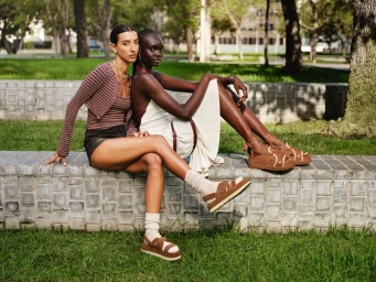 Two women sitting on a low tiled retaining wall outdoors, wearing brown platform sandals, knit socks, and casual apparel.